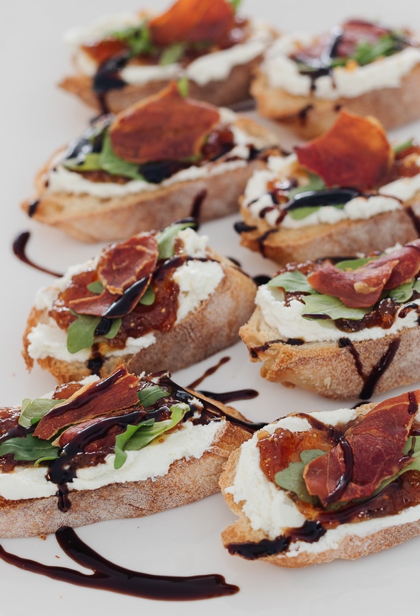 toasted baguette topped with goat cheese crispy prosciutto and arugula on a white platter drizzled with balsamic vinegar