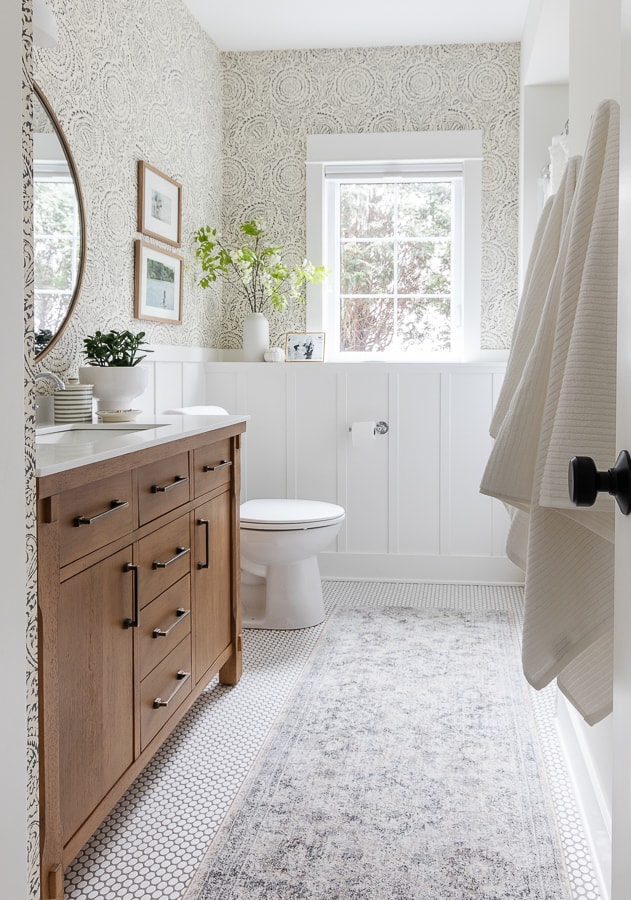 white bathroom with penny tile and wallpaper with wood vanity